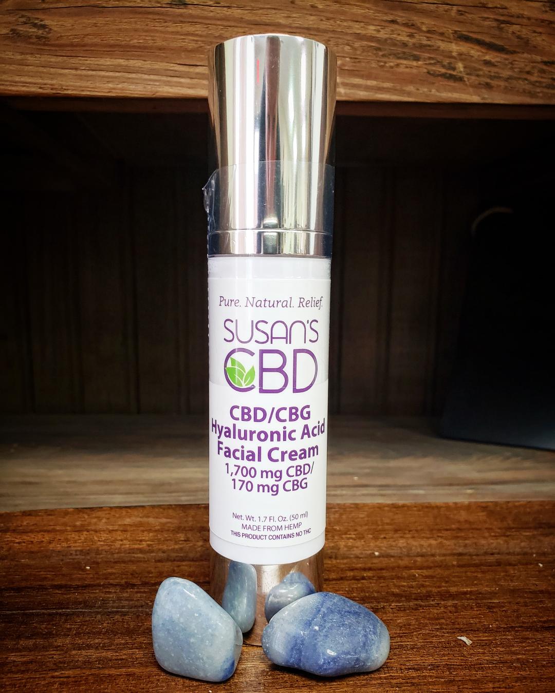 A bottle of Susan's CBD/CBG Hyaluronic Acid Facial Cream displayed on a wooden surface with two blue stones in front of it.