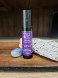 A roll-on bottle labeled 'Susan's CBD/CBG Calming Roll On' with a dark liquid visible inside, placed next to a smudge stick and a crystal on a wooden surface.