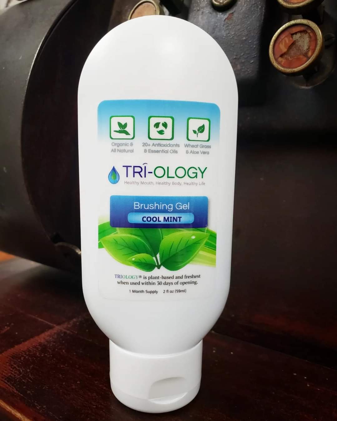 A tube of Triology Brushing Gel with cool mint flavor, placed on a surface with a background that includes a register and other vintage items. The packaging is predominantly white with green and blue accents, and text that highlights the product's natural ingredients and benefits.