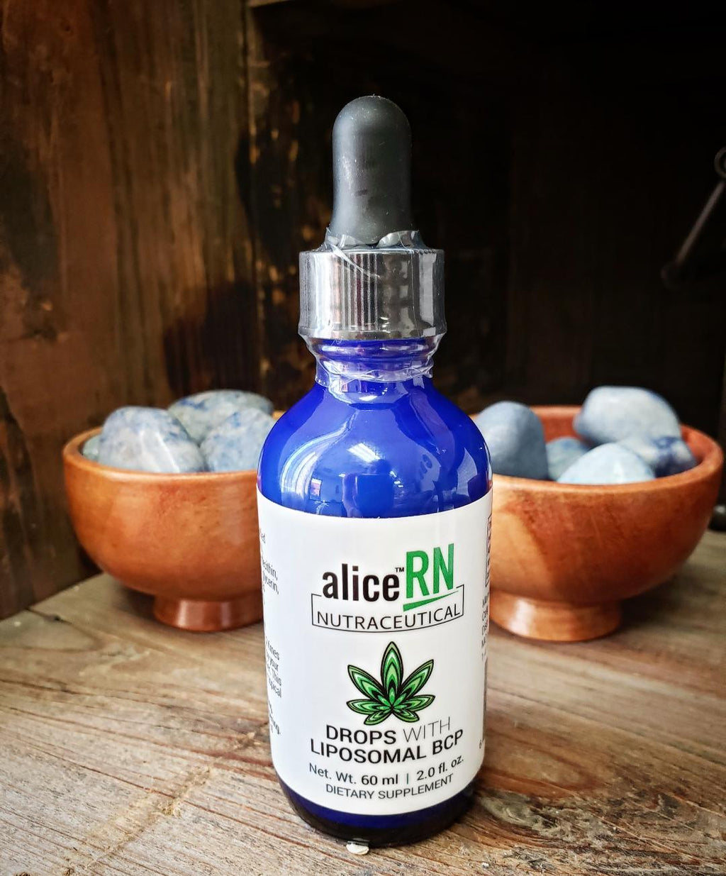This image features a bottle of Alice BCP Liposomal Drops, prominently displayed on a wooden surface with a rustic backdrop. The bottle is a striking blue color, which draws attention, and it has a clear dropper for easy application.